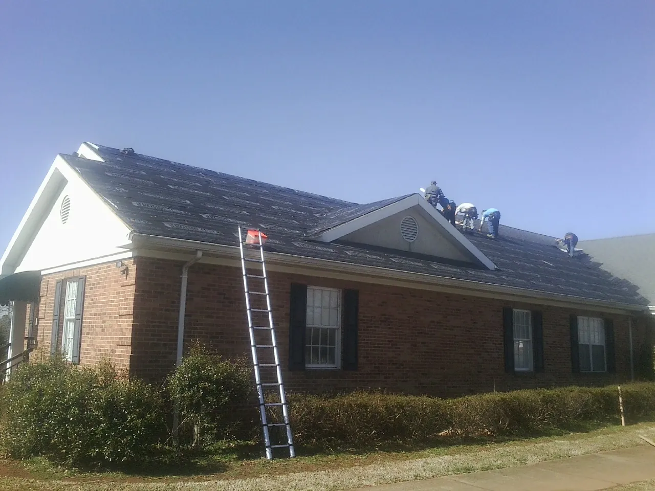 Crew tearing shingles off a brick ranch roof with a ladder set — Metal Roof Repair work by our crew in Acton