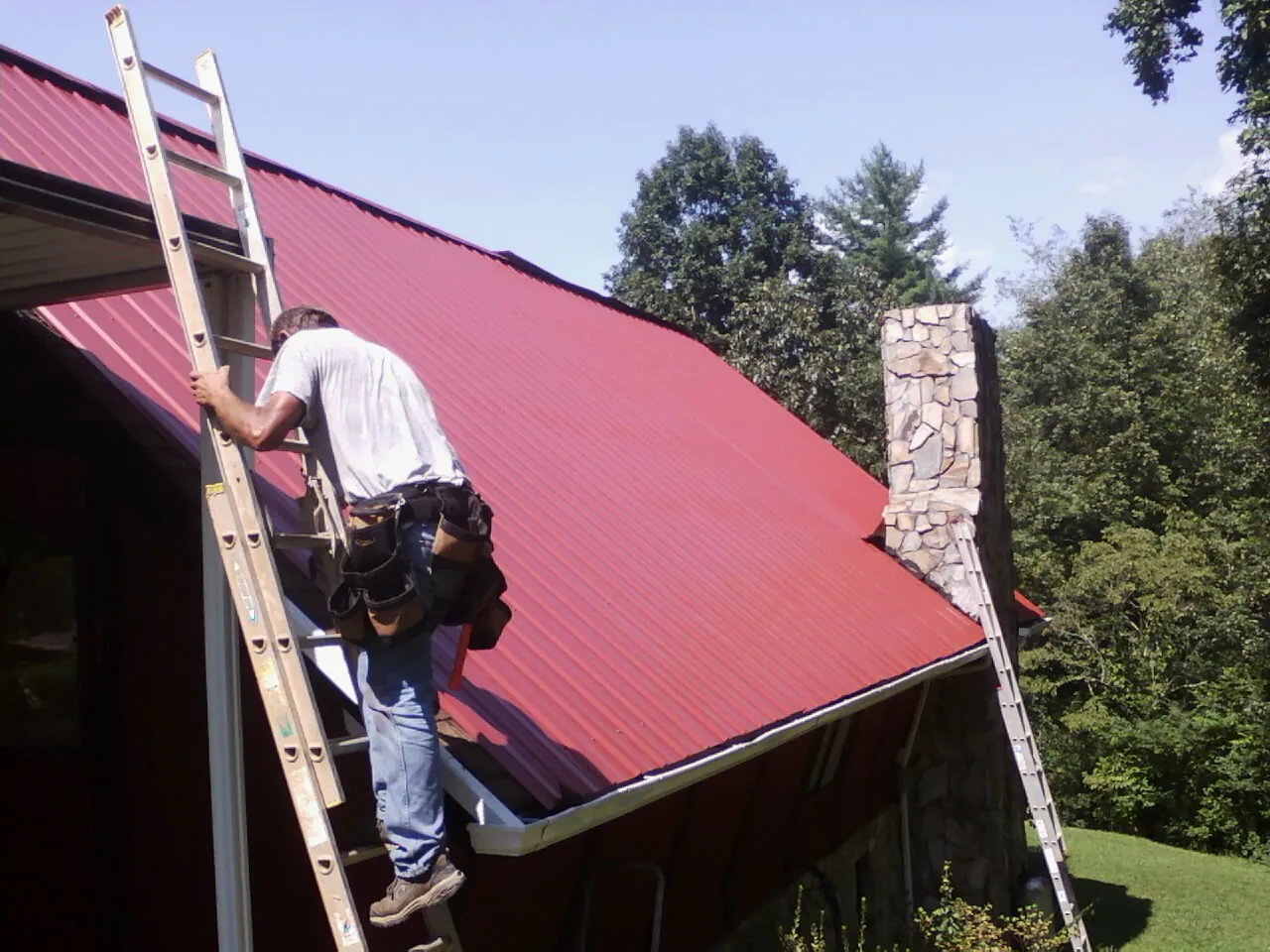 Roofer on a ladder beside a red metal roof and stone chimney — Soffit & Fascia Repair by Roofing Brothers in Acton